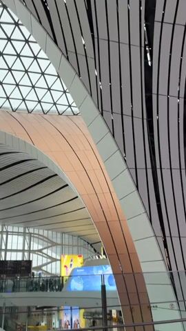 The video pans across the interior of a modern airport terminal. The ceiling is a prominent feature, with a large, geodesic dome structure and undulating, ribbed panels in white and black, accented with bronze-colored metallic sheeting. Natural light streams through the triangular glass panels of the dome. Below, the polished floor reflects the architectural elements. Several signs are visible, including directional signs with Chinese characters and a sign indicating "4F." People are seen walking, some with luggage, and others are at check-in counters. A large digital display screen is visible in the background. The overall impression is of a spacious and architecturally striking airport environment.