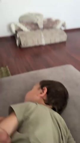 A young child wearing a light green t-shirt is filmed indoors. The child's head is turned to the left, then moves closer to the camera and faces forward. The child has dark hair and appears to be looking at the camera with a concerned expression. The child's right hand is placed over their chest. In the background, rolled-up rugs are stacked against a wall, and wooden flooring is visible. The lighting suggests an indoor environment.