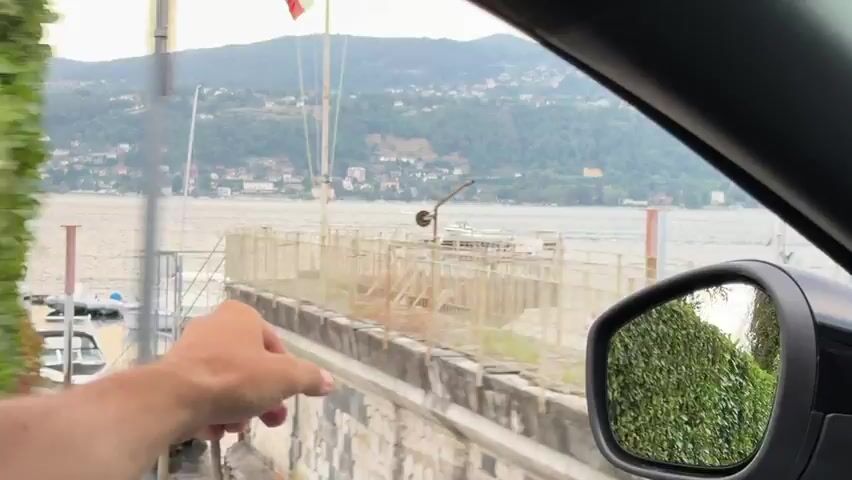 The video begins from the perspective of someone inside a car, looking out the passenger side window. A silver Mini Cooper passes by. The view then shifts to a red SUV. Next, a hand is seen pointing towards a table and benches by the water. The camera then moves to show a wider view of a lake with hills and houses in the background. A boat is visible on the water. The hand is seen again, gesturing towards the water and the distant shore. The video continues to show the lake and the hillside town, with the hand occasionally appearing in the foreground. The scene appears to be a scenic lakeside location with boats on the water and a town built on the hills.