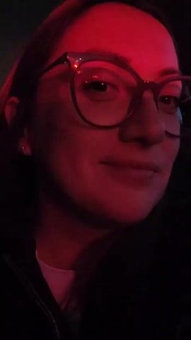 The video shows a close-up of a person's face illuminated by a red light. The person is wearing glasses and has glitter makeup on their cheek. They are wearing a dark jacket and a white shirt underneath. The background is dark. The person is looking at the camera and appears to be smiling slightly.