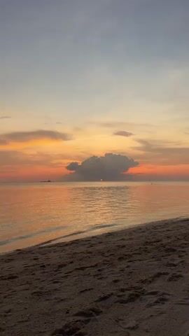 A still, wide shot reveals a beach scene at sunset. The sky is a gradient of orange, yellow, and grey, with a large, dark cloud obscuring most of the setting sun, leaving only a sliver of red visible. The ocean reflects the sky's colors, shimmering with orange hues. Gentle waves lap against the sandy beach, which is visible in the foreground with patterns from footprints or other disturbances. No people are visible in the shot.