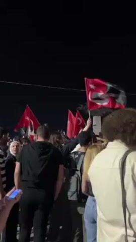 Here is a concise description of the video:

The video shows a crowd of people gathered at night, waving Turkish flags. Most of the individuals are facing the same direction. A few people are holding their phones, seemingly filming or taking photos. The location seems to be a public square, as evidenced by the open space and what looks like a monument or statue in the distance. The night sky is dark, with a street light visible above the crowd. The mood appears to be celebratory or patriotic.
