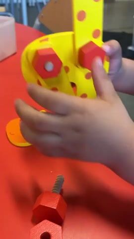A child is playing with a wooden toy giraffe on a red table. The toy is yellow with red polka dots and has red hexagonal pieces attached. The child is using their hands to manipulate the toy, seemingly trying to assemble or disassemble it. One piece of the giraffe head is lying separately on the table, along with a wooden screw. The background shows a classroom setting with chairs and tables. A person walks in the background.