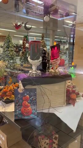 The video begins with a close-up of a Halloween-themed display in a store window. The display features large decorative candles, pumpkins, faux autumn leaves, and a whimsical painting of pumpkins. In the background, Christmas trees are visible, suggesting a transition between holidays or a store that sells items for multiple occasions. The camera then pans to the right, revealing the interior of what appears to be an empty or sparsely populated shopping mall. The flooring is tiled, and there are large pillars with a faux wood design. In the distance, stores with closed gates are visible. The overall impression is one of a quiet, possibly disused commercial space. There are no people visible in the video.