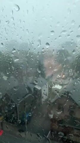 The video shows a view through a window covered in raindrops. The raindrops are varied in size and shape, distorting the view of the scene outside. The scene outside appears to be a suburban or urban environment with houses and trees. The weather is overcast and rainy, with a general grayness to the scene. Lights from vehicles or buildings are visible in the distance, blurred by the rain. The camera perspective is static, looking out from an interior space.