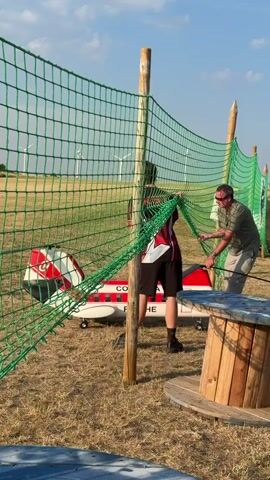 The video shows two men setting up a green net that is attached to wooden posts. The net is strung across a grassy area, with a large wooden spool visible in the foreground. In the background, a small, red and white airplane with "COLUMBIA" written on its side is on the ground. Several wind turbines are visible in the distance, along with a clear sky and some fluffy clouds. Other people and tents are also present further in the background, suggesting an outdoor event or gathering. The men appear to be working together to adjust the tension or position of the net.