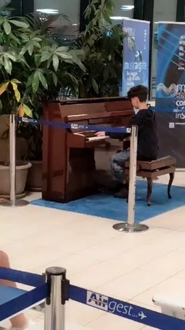 A young person with dark, curly hair, wearing a black hooded sweatshirt and distressed denim jeans, sits on a wooden piano bench playing an upright piano. The piano is dark brown and appears to be a grand piano. The bench has ornate legs. The piano is situated on a blue rug in what appears to be a public space, possibly an airport terminal, given the signs in the background. One sign is blue and white with text that is difficult to read but includes the words "musical gate" and "concerto." Another sign is partially visible to the right. A tall potted plant with large green leaves is to the left of the piano. Metal stanchions with blue tape are set up around the piano area, creating a boundary. The floor is tiled in a light color. The lighting appears to be artificial, possibly from overhead fluorescent lights. The person is facing away from the camera, focused on playing the piano. The scene is captured from a medium distance, with the focus on the person and the piano.