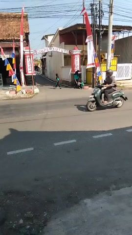 The video shows a street scene with several people near an archway. A yellow truck drives past. There are Indonesian flags and decorative banners displayed. Two children are visible near the archway, and two adults are seated or leaning near it. The street is paved with asphalt and has white lane markings. Buildings and trees are visible in the background.