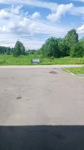 The video shows an outdoor scene on a cloudy day with a bright sky. In the foreground, there is a gray asphalt surface with some dark stains. In the middle ground, there is a road or pathway leading into a grassy area with trees and bushes. There is a small white structure with a blue roof in the distance, possibly a small shed or shelter. The background consists of more trees and a line of power lines overhead. The overall environment appears to be rural or semi-rural. There are no people or animals visible in the video.