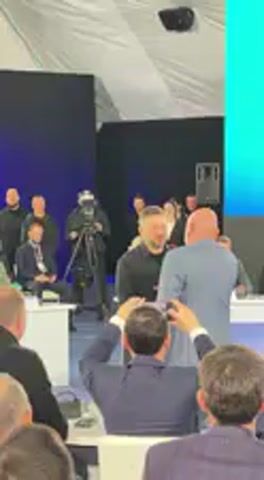 The video captures an indoor event, likely a ceremony or official gathering, set in Barvinok, Ukraine. The scene features a central interaction between two men. One man, wearing a black long-sleeved shirt and sporting a beard, is receiving a small, rectangular item wrapped in red/purple paper. He smiles broadly. The other man, who is bald with a beard and dressed in a light blue suit jacket over a black shirt with a white lanyard, is handing over the item and also smiling.

In the foreground, the back of a person's head is visible, holding a smartphone up, seemingly recording the event. Another individual operating a professional camera on a tripod can be seen in the background to the left. Several other attendees are scattered throughout the background, some standing, some seated at white tables. The setting includes dark blue backdrops or curtains and a large, brightly lit turquoise screen on the right side of the frame. The audio features a male voice speaking in Ukrainian, announcing names such as "Володимиру Івановичу" (Volodymyr Ivanovych) and mentioning "сільського голови" (village head/mayor), suggesting an official presentation or award. The overall atmosphere appears positive and celebratory.