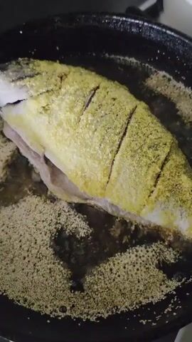 A whole fish, coated in a yellow spice mix, is frying in a black skillet filled with bubbling oil. The fish is scored with three diagonal cuts across its back. The skillet appears to be on a stovetop, and the background is dark and out of focus. The video shows the fish from a close-up, top-down perspective.