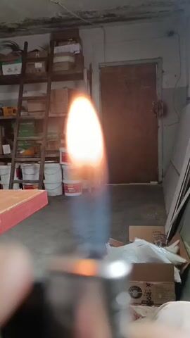 A close-up shot focuses on a lighter igniting a flame, with the flame dominating the frame as it flares upwards. The background is a dimly lit interior, appearing to be a workshop or storage area. Shelves stacked with boxes, buckets, and a ladder are visible on the left. To the right, a wooden door and more storage items can be seen. The ground is concrete. The lighting suggests it could be any time of day, but the interior setting doesn't provide clear weather cues. No people are clearly visible, though a hand is out of focus in the foreground, holding the lighter. The overall impression is of an industrial or utility space, with the central activity being the controlled ignition of a flame. Some text is visible on the buckets and boxes in the background, but it is too small and blurry to read.