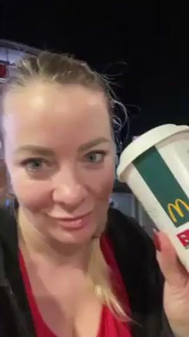 A woman with blue eyes and blonde hair pulled back into a braid is standing outside at night. She is wearing a red shirt and a black jacket. She holds a large paper cup with a white lid, decorated with a McDonald’s logo and then a beer advertisement. She rotates the cup to show the design, then takes a drink from it. The background is dark, with some lights visible.
