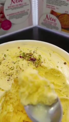 The media file shows a close-up shot of a bowl of yellow ice cream, possibly mango-flavored, topped with chopped pistachios and some reddish-pink dried rose petals. A spoon is in the process of scooping out a portion of the ice cream, with a generous amount already on the spoon, glistening as it melts slightly. The ice cream appears creamy and dense.

In the background, out of focus, are signs or menu boards. One sign has the word "New" prominently displayed in a pink font within a circular design. Another sign to the right also features "New" and what appears to be an image of a dessert, possibly a tart or pie with passionfruit. Some prices are visible on the signs, such as "€23.00". The overall setting suggests an ice cream parlor or dessert shop. The lighting is bright, indicating it's likely daytime, and the overall mood is inviting and indulgent, focusing on the appealing texture and toppings of the ice cream. There are no people visible in the frame. The location context is Bedford, United Kingdom.