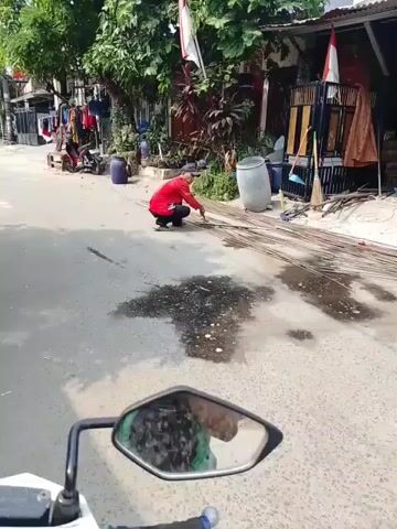 The video shows a person wearing a red shirt and dark pants crouching on the side of a paved road. They appear to be working with long, thin metal rods laid out on the road. The environment looks like a residential street, with houses and some vegetation visible in the background. Laundry is hanging on a line outside one of the houses. A motorcycle's side mirror is in the foreground, reflecting the scene. There are some dark, wet patches on the road in front of the person.