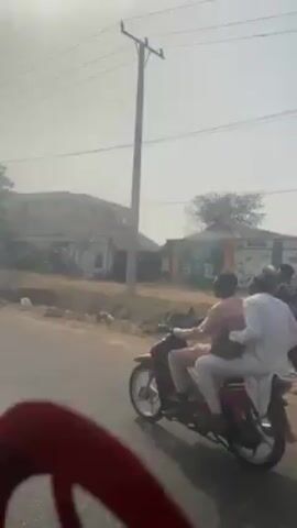 A car is driving down a road, passing a couple of motorcycles. The sky is very bright and hazy. The road is paved and in good condition, and there are buildings and trees alongside the road. A red car seat can be seen in the foreground of the video, probably where the video camera is sitting.