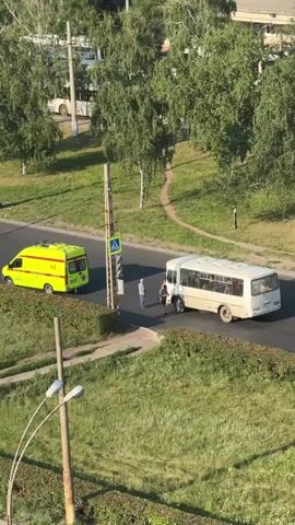 The video depicts an outdoor scene, likely in Tolyatti, Russia, during the daytime, possibly late afternoon or early evening due to the warm, golden light and long shadows. The setting appears to be a suburban or semi-urban area with a road, a grassy verge, and trees.

The main subjects are a yellow ambulance vehicle and a white bus. The ambulance is positioned on the left side of the road, facing away from the camera, and its hazard lights are illuminated, indicating it might be responding to an emergency or is parked due to an incident. Visible text on the ambulance includes "03" and "МЕДИЦИНСКАЯ ПОМОЩЬ" (Medical Assistance), along with a red cross emblem.

On the right side of the road, near a pedestrian crossing with a traffic light, is the white bus. Two people are standing near the bus, appearing to be in conversation or observing the situation. One person is wearing a blue shirt and jeans, and the other is wearing a light-colored dress and appears to be holding a phone. A third person, wearing dark clothing and a backpack, is standing closer to the bus. The bus has windows, and some passengers can be seen inside, though their faces are not discernible. The bus also has some Cyrillic text on its side, though it's largely illegible due to the distance and resolution.

The activity suggested is a potential traffic incident or a medical emergency response. The presence of the ambulance and the people gathered near the bus on the road points to an event that has caused a stop or a need for assistance. The overall mood seems calm, with no overt signs of distress, but the circumstances suggest a non-routine situation. The weather appears to be clear and pleasant, with lush green grass and full trees indicating a warm season.
