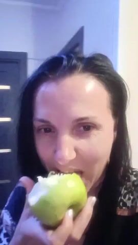 This media file is a short video clip featuring two people. The primary subject is a woman in the foreground, who is eating a green apple. She has dark hair and is looking towards the camera. To her left and slightly behind her, a man is visible. He has light-colored hair and appears to be smiling. He is topless, showing a portion of his chest. The background suggests an indoor setting, possibly a room in a home. There is a dark doorway or piece of furniture visible behind the subjects. The lighting is somewhat dim, indicating it could be evening or indoors without bright illumination. The woman's expression is focused on eating the apple, while the man's expression is cheerful. The context provided indicates the location is Penza, Russia, which offers no visual cues in the video itself but is important for understanding the setting. No text is visible in the video.