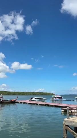 The video shows a bright, sunny day with a clear blue sky filled with scattered white clouds. The scene is a body of water, likely the ocean, with a wooden pier extending into it. On the pier, there are at least two speedboats docked. One is white with blue and black accents, and appears to be labeled "S.Y. CHANGING TOUR". The other speedboat is docked further out on the pier and is decorated with bright pink and blue patterns. In the distance, a lush green tree line is visible, suggesting land or an island. The water is calm with gentle ripples. In the sky, a bird is seen flying. The overall impression is a serene and idyllic tropical setting.