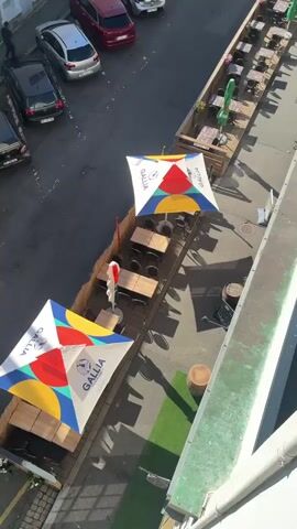 The video pans up from a small parking area with outdoor seating in front of a white building. The outdoor seating has umbrellas with a Gallia logo. The parking lot has several cars. People are walking on the street and entering the building. The video moves up to show the surrounding city, with buildings of various heights and architecture. The sky is blue with some clouds. The video appears to be taken from a high vantage point, possibly a balcony or upper floor of a building.