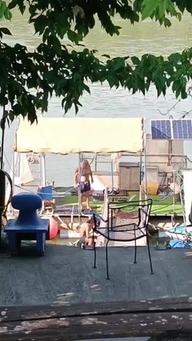 A video shows a man climbing out of the water onto a boat. The man wears swim trunks and appears to be rinsing himself off with a hose. Another individual is partially visible in the water near the boat. The boat has solar panels on top and a yellow awning. The scene is viewed from behind some foliage, with a body of water visible in the background.