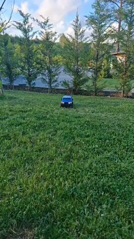 A blue remote-controlled toy SUV is shown driving on a grassy lawn. The toy truck has a spare tire mounted on the back and a roof rack. In the background, there are several tall, thin trees and a house. The sky is blue with some clouds. The camera appears to be held by someone walking behind the toy truck, sometimes showing the truck in front, sometimes showing the truck from above as it moves away.