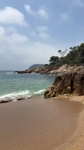 A video shows a beach with sandy shores, rocky cliffs, and green trees. The ocean waves are gently rolling onto the shore. The sky is partly cloudy and the weather appears to be fair. The scene is peaceful and serene.