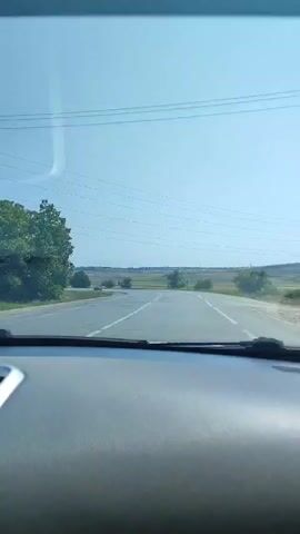 The video shows a point-of-view shot from inside a car traveling on a paved road. The road has white lines marking the lanes. On the left side of the road, there are trees and fields. On the right side, there are also fields and some trees. The sky is clear and blue. There are power lines visible in the sky. There are other cars on the road, with one car visible in front and another passing in the distance on the left. The dashboard of the car is visible in the foreground.