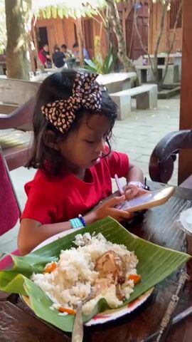 A young girl wearing a red t-shirt and a patterned hair bow sits at a wooden table, holding a smartphone and looking at its screen. In front of her is a plate of food, consisting of white rice and a piece of fried poultry, served on a banana leaf. An adult's arm reaches into the frame, placing a "RESERVASI" sign on the table. The camera then pans across the table, showing two plates of food, each with rice, a piece of fried poultry, and red chili sauce on a banana leaf. Two plastic cups with drinks, one yellow and one clear with a dark liquid, are also on the table. The background shows a restaurant or cafe setting with wooden structures and other patrons.