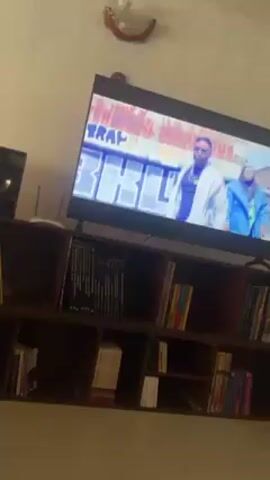 The video shows a television screen with a music video playing. The music video features people and a monster truck. The television is sitting on top of a bookshelf filled with books. The camera angle is slightly low, looking up at the screen.