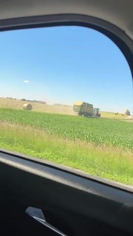 This video shows a rural landscape from the perspective of someone inside a vehicle, looking out a window. The scene is a bright, sunny day with a clear blue sky. The foreground is dominated by the interior of the car, with the dark frame of the window in the foreground and a glimpse of a car door handle.

The main subject of the video is a large agricultural field. In the middle ground, a green tractor is pulling a yellow trailer, kicking up a significant amount of dust as it moves across the field. The field itself is a mix of tall green crops, likely corn, and dried, harvested grass. In the background, there are several large, round hay bales scattered in the field. Further in the distance, another piece of agricultural machinery, possibly a baler, is visible. The overall setting is a rural farming area, with fields stretching out towards the horizon.

The activity taking place is agricultural harvesting, likely of hay or silage, indicated by the presence of the tractor, trailer, and hay bales. The dust cloud suggests the machinery is actively working. The time of day appears to be midday, given the bright sunlight and clear sky. The weather is warm and dry, conducive to fieldwork. The location cues, such as the style of the agricultural machinery and the types of crops, combined with the provided location context of Tuntenhausen, Germany, suggest this is a farm in Bavaria. There are no people prominently visible in the footage. The overall mood is one of industrious agricultural activity on a beautiful summer day. No text is visible in the video.