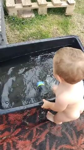 A young child, wearing only a diaper, sits on the edge of a black plastic tub filled with water. The child is reaching into the water with their right hand, playing with a small toy that is mostly submerged. The toy appears to be blue and green. The child's left hand is resting on the edge of the tub. The tub is placed on a red and black patterned surface. In the background, there is green grass and wooden pallets. The scene appears to be outdoors.