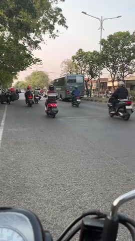 The video shows a street scene with multiple motorcycles and a bus. The perspective is from someone riding a motorcycle, looking forward. Several motorcycles are in motion on the road, some with riders wearing helmets. One rider in the foreground is wearing a yellow jacket and helmet. Trees line the side of the road, and buildings are visible in the background. The sky appears overcast. The scene depicts traffic on a road.