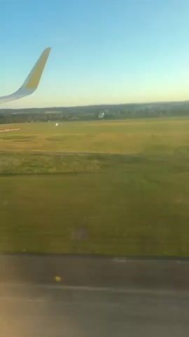 The video shows a view from an airplane window as it taxis on a runway. The airplane's wing is visible on the left side of the frame, with its wingtip pointing upwards. The runway is made of asphalt and has white markings. Beyond the runway, there is a large grassy field with some scattered buildings and trees in the distance. The sky is clear and blue, with the sun setting, casting a warm golden light on the landscape. The plane appears to be moving at a slow speed. There are no visible people or passengers in the frame.