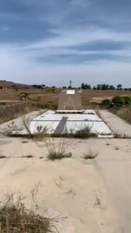 The video shows an elevated, weathered concrete structure with a white tile design, likely a mausoleum or monument, in a sparse, dry landscape under a partly cloudy sky. The structure has some weeds and cracked tiles. In the background, the landscape consists of dry grasses and scattered trees. A cross stands at the head of the elevated structure.