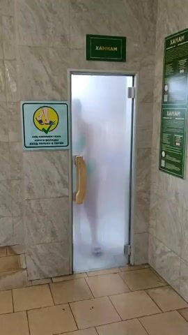 A video shows a person opening a glass door into a steamy room labeled "ХАММАМ" in Russian. The person, wearing a green bikini, is partially obscured by steam. Inside the room, a young boy in blue swim trunks stands, also surrounded by steam. The room appears to be a hammam or steam room. There are signs on the exterior wall; one shows a pictogram of feet with an arrow indicating "no shoes," and text below it in Russian reads, "Вход в хамам без обуви" which translates to "Entrance to the hammam without shoes." Another sign to the right is partially visible and appears to contain text and a QR code. The overall impression is of a steamy, enclosed environment, likely a spa or bathing facility.