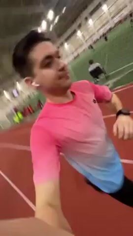 Here is a description of the video:
The video shows a man running on an indoor track. The man is wearing a pink and blue gradient shirt, black shorts, and wireless earbuds. He is holding the camera in his hand, capturing a selfie view as he runs. Other people can be seen in the background, some running on the track and others on the green field in the center. The environment is a large indoor sports facility with a high ceiling and artificial lighting. The track is red, and there are multiple lanes. The video appears to be taken during a workout or casual run.