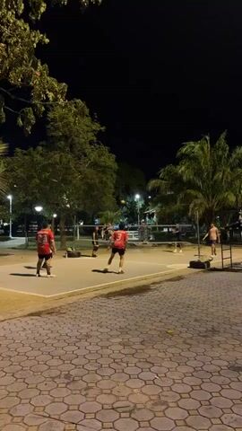 At night, a group of people are playing a game on a concrete court surrounded by trees and palm trees. The court is lit by streetlights, and a net is strung across the middle. Several people are positioned on each side of the net, wearing casual clothing. One person is wearing a red shirt with a white number. The camera is static and captures the scene from a distance, showing the players in motion as they interact with a ball. The environment includes paved walkways and the night sky above.