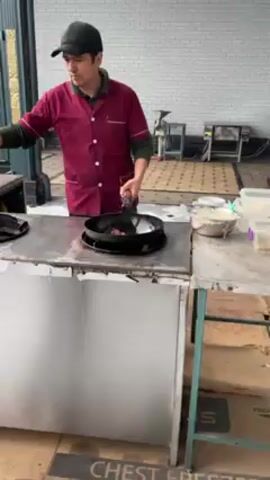 A man in a red chef's coat and a black baseball cap is stir-frying food in a wok. He’s standing behind a stainless steel cooking station. As he tosses the ingredients with a spatula, a large flame erupts from the wok. The cooking station is surrounded by other kitchen equipment and supplies, and a brick wall is visible in the background. The floor is tiled in a decorative pattern.