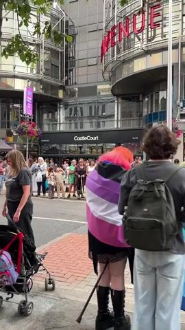 The video shows a street scene, likely during a parade or festival. In the foreground, a person with blonde hair wearing a grey t-shirt and black pants stands facing left, with a stroller and a pink and beige jacket beside them. To the right, a person's back is visible, holding a flag with purple, pink, white, and blue stripes, suggesting an LGBTQ+ pride event. A group of people stand across the street, watching something happening in the distance. Behind them, a multi-story building with glass windows and signage for "CastleCourt" and "New Look" is visible. There are banners and hanging flower baskets on the building. Further down the street, more people are gathered, and a colorful umbrella can be seen. The overall atmosphere appears festive and crowded.