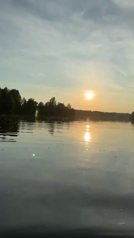 A video captures a sunset over a tranquil body of water, with the foreground dominated by a spray of water droplets. The sun, a bright orb, is partially obscured by clouds and casts a shimmering reflection on the water's surface. A treeline borders the horizon. The overall scene conveys a sense of peacefulness and natural beauty.