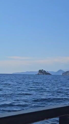 A boat travels across a blue sea. In the distance, there are islands and mountains. The sky is a clear blue.