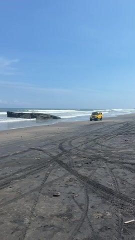 The video shows a yellow Land Rover Defender parked on a black sand beach under a clear, blue sky with scattered clouds. Tire tracks crisscross the foreground sand. Waves break on the shore in the background, near a rocky outcrop. The scene is static with no visible people or actions taking place.