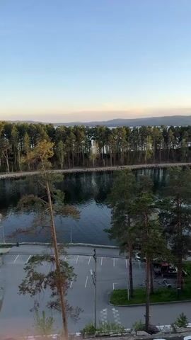 The camera pans from left to right, revealing an elevated view of a body of water with a distant shoreline. A dense line of evergreen trees separates the water from the view. The sky above is a clear gradient of light blue transitioning to a warmer yellow near the horizon. Below the camera's vantage point is a paved parking area with a few parked cars, including a blue one. Tall pine trees dot the landscape near the parking lot and waterfront. Beyond the trees, across the water, a hazy mountain range is visible in the distance. The overall scene suggests a calm, serene environment with natural beauty.