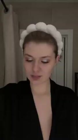 A young woman wearing a black robe and a white, puffy headband stands in a bathroom and holds up a pink and a green toothbrush. She makes a series of facial expressions. There is a shower curtain to the left and a white door behind her.