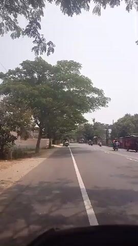 The video shows a first-person perspective of a person riding a vehicle, likely a scooter or motorcycle, on a paved road. The road has a white line dividing the lanes. Trees with green leaves line the left side of the road, and there are buildings visible. On the right side of the road, there are more trees and some structures that appear to be shops or houses. Traffic is present on the road, including other motorcycles and cars, and at least one red bus is visible in the distance. The sky is bright, indicating daytime. The rider's handlebars or dashboard are partially visible in the foreground. The overall scene depicts a typical roadside environment in a moderately populated area, possibly in a tropical or subtropical region due to the dense foliage.
