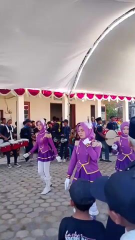 A group of young women in matching purple and gold marching band uniforms with white boots are performing a dance routine under a white tent. They are wearing purple hijabs. In the background, a marching band is playing drums. There are spectators watching the performance.