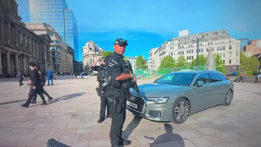 The video shows two police officers standing in a paved public square. Both officers are wearing tactical gear, including vests and helmets. The officer in the foreground is holding a rifle. The officers are standing next to a silver Audi station wagon. In the background, there are several buildings, including a modern high-rise building and older, more ornate structures. There are also a few other people walking in the square and sitting on benches. The sky is blue with some clouds. The lighting suggests it is daytime, likely morning or afternoon.