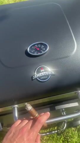 A person holds a cigar in their left hand in front of a black Jamestown Grill with a temperature gauge reading 225 degrees Fahrenheit. The person opens the grill, revealing several items cooking on the grates, including two pieces of meat, several pieces of foil-wrapped food, and smoke rising. The grill is sitting on grass.