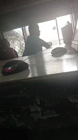 The video shows the interior of a dimly lit room, likely an office or shop.  Two people are visible, their faces indistinct due to backlighting from a bright window behind them. They appear to be sitting at a counter. A laptop is on the counter, along with a computer mouse with a red light illuminated. The counter itself is painted white and has a textured, possibly aged or worn surface. Wires and electrical fixtures are visible on the ceiling.